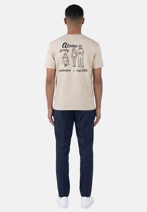 Uomo che indossa una maglietta beige con disegno lineare nero di tre persone e testo "Atomo factory petanque cup 1954" sulla schiena, pantaloni blu navy, scarpe bianche.