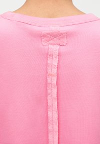 Κοντινό πλάνο της πίσω πλευράς ενός ανοιχτό ροζ πουκαμίσου που δείχνει μια κάθετη λωρίδα υφάσματος ραμμένη από το λαιμό μέχρι το πάνω μέρος της πλάτης, αναδεικνύοντας την υφή και τις λεπτομέρειες της ραφής.