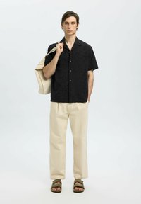 Chemise noire à manches courtes avec motif texturé, pantalon large beige, et sandales vertes. Modèle tenant un sac fourre-tout beige.