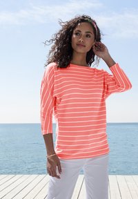 Koralfarbener, gestreifter, langärmeliger Strickoberteil mit weitem Ausschnitt und lockerer Passform, kombiniert mit weißen Hosen. Das Model streicht sich die Haare vor einer Strandkulisse.