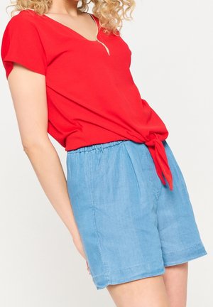 Vrouw draagt een rood kort mouwenshirt dat in de taille is gestrikt en lichtblauwe, hooggetailleerde shorts tegen een effen achtergrond.