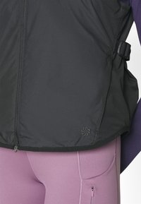Svart zip-up jacka med texturerad tyg. Har en sidospänne och en liten logotyp vid fållen. Matchad med figursydda ljuslila leggings.