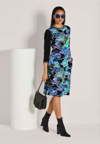 Vrouw in een blauwe bloemenjurk, zwarte enkellaarsjes, blauwe zonnebril, met een zwarte handtas, staand naast een hoge witte cilindrische voet.