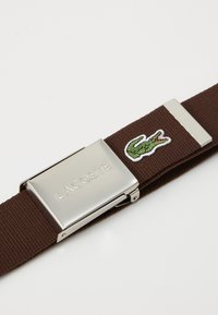 Obdĺžnikový hnedý tkaný opasok so striebornou kovovou prackou. Na pásiku sa nachádza logo Lacoste a výšivka zeleného krokodíla.