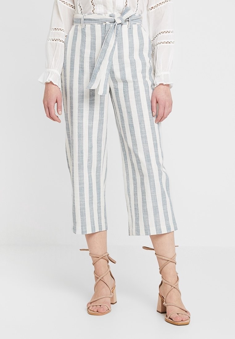 Gestreepte cropped pantalons in lichtblauw en witte linnenmix, met een ceintuur in de taille en een wijd beenontwerp. Gecombineerd met beige sandalen met hakken en bandjes.
