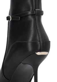Black leather ankle boot with a pointed toe, a sleek silhouette, a thin high heel, and a gold accent near the heel. Features a strap detail.