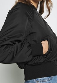 Czarna bomberka z błyszczącej, gładkiej tekstury, z żebrowanymi mankietami i dołem oraz bocznymi kieszeniami. Prosty design, dopasowany krój podkreślający talię.