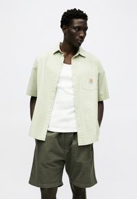 Άνδρας που φοράει κοντομάνικο πουκάμισο σε ανοιχτό πράσινο με υφή πάνω από λευκή φανέλα και χακί βερμούδα, στέκεται με τα χέρια στις τσέπες.