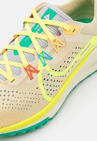 Chaussure de trail Nike beige avec tige perforée, lacets jaunes, œillets colorés, semelle verte et logo Swoosh accentué de jaune.