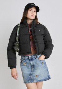 Svart puffjakke med høy krage, glidelås foran og elastiske mansjetter; kombinert med et lyseblått denimskjørt med broderte detaljer.