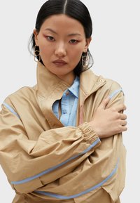 Beige lightweight jacket with blue accents and a high collar. Features elastic cuffs and a front zipper. Underneath is a light blue button-up shirt.