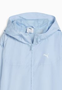 Veste coupe-vent Puma bleu clair avec une capuche doublée en mesh, fermeture éclair complète et un logo discret sur la poitrine. Texture lisse, design léger.