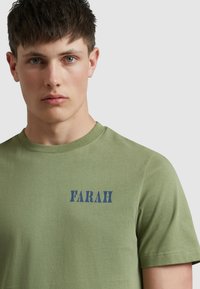 Koszulka z krótkim rękawem w kolorze oliwkowym, wykonana z bawełny, z granatowym logo "FARAH" na lewej piersi. Okrągły dekolt i gładka tekstura.