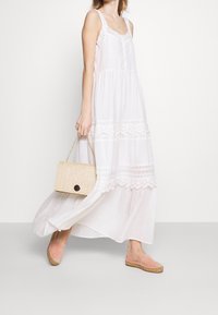 Hvid bomulds maxi kjole med blonder, runde knapper og lagdelte design. Holder en vævet beige håndtaske med en sort lukning, parret med lyserøde espadrilles.