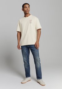 Beige katoenen t-shirt met korte mouwen, met de opdruk "Road to Freedom" in bruin. Geslaagd met blauwe denim jeans en witte sneakers.