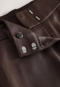 Pantalon marron foncé avec ceinture élastique, braguette zippée, fermeture par bouton et deux crochets métalliques sur la ceinture.