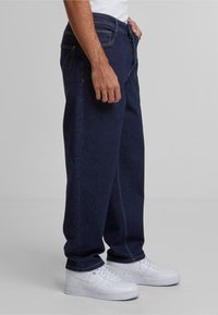 Jeans de mezclilla azul oscuro con un corte holgado, que cuentan con costuras amarillas, cinco bolsillos y un cierre de cremallera, combinados con zapatillas blancas.