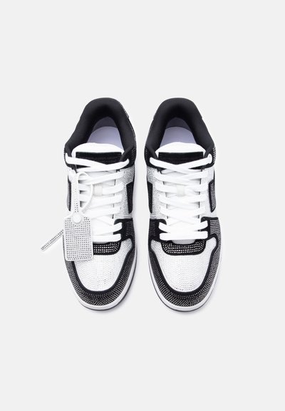 Ζευγάρι ασπρόμαυρα αθλητικά παπούτσια με λευκά κορδόνια και μια λαμπερή ορθογώνια ετικέτα δεμένη στο αριστερό παπούτσι, σε λευκό φόντο.