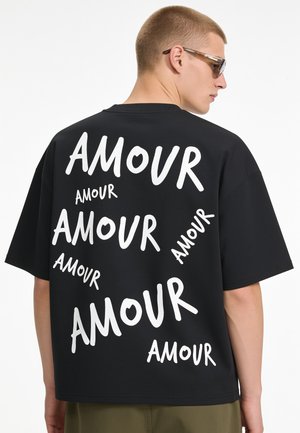 Jeune homme portant des lunettes de soleil et un t-shirt noir oversize avec le mot "AMOUR" imprimé plusieurs fois en blanc dans le dos.