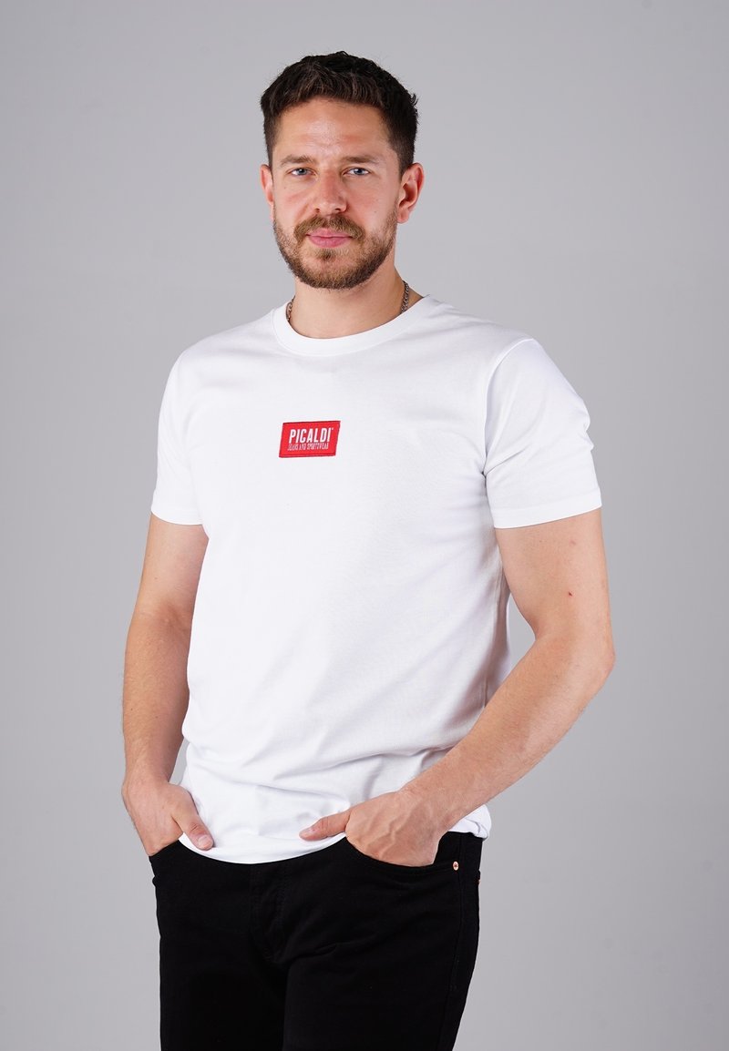 Mann mit kurzem dunklem Haar und Bart, der ein weißes T-Shirt mit rotem Logo und schwarze Hosen trägt, mit den Händen in den Taschen, vor einem grauen Hintergrund stehend.
