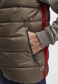 Padded Weste in olivgrün mit einem roten Streifen an der Seite. Verfügt über Reißverschlusstaschen und Schnürhardware mit roten und weißen Akzenten.