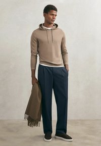 Mand iført beige hættetrøje, hvid undertrøje, navybukser og sorte sneakers, der holder et brunt frynset tørklæde i den ene hånd.