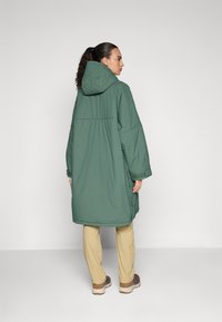 Abrigo largo y verde acolchado con capucha, que presenta un corte relajado, puños elásticos y detalles acolchados. Usado sobre pantalones beige y zapatos.