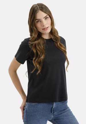 Młoda kobieta z długimi falowanymi włosami, ubrana w prostą czarną koszulkę i niebieskie dżinsy, z rękami za plecami, patrząca na kamerę.