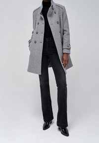 Trench coat grigio a spina di pesce con design doppiopetto, chiusure lampo frontali e grandi bottoni, abbinato a jeans neri a vita alta a palazzo e stivali.