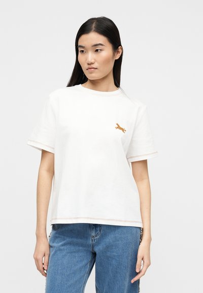 Νεαρή γυναίκα με ίσια μαύρα μαλλιά, που φοράει ένα λευκό T-shirt με ένα μικρό κεντημένο τίγρη και μπλε τζιν, στέκεται μπροστά σε απλό φόντο.