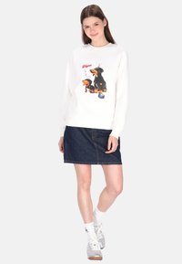 Weißes Sweatshirt mit einem bunten Motiv eines Hundes und einer Cartoonfigur, kombiniert mit einem dunklen Jeansrock und grauen Turnschuhen.