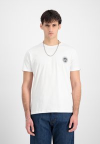 Hvid bomulds T-shirt med rund halsudskæring, korte ærmer og en frontlomme med et cirkulært sort grafisk design. Kombination med blå denimjeans.