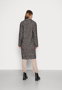 Cappotto lungo a motivi in tessuto nero e grigio, con taglio dritto, maniche lunghe e colletto a lancia, abbinato a pantaloni beige e stivali neri.