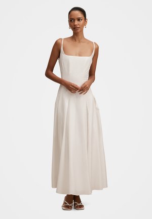 Kvinde stående, iført en ærmeløs, cremefarvet kjole til anklerne med tynde stropper og plisseret nederdel, kombineret med hvide hælede sandaler.