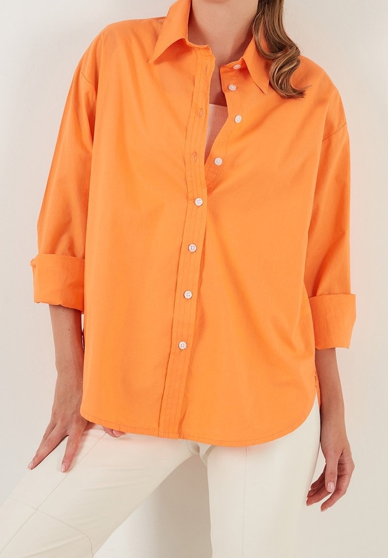 Orange skjorta i lätt tyg med krage, långa ärmar uppvikta och en rundad nederkant med vita knappar.