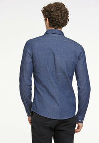 Camicia in denim blu scuro con colletto classico, maniche lunghe e polsini con bottoni. Texture liscia con una leggera lucentezza e orlo curvo.