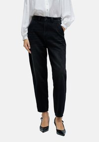 Jeans negros de cintura alta con pierna ajustada, diseño clásico de cinco bolsillos y ligero desgaste. Combinados con una camisa blanca de manga larga.