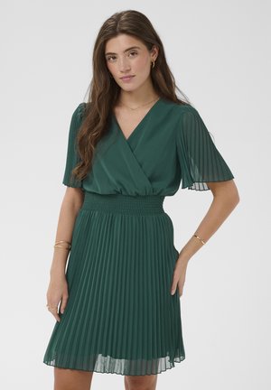 Młoda kobieta o długich brązowych włosach, ubrana w ciemnozieloną plisowaną sukienkę z krótkimi rękawami, złote kolczyki koła oraz złote bransoletki.