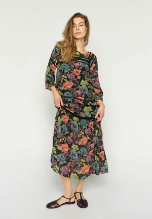 Pikkade juustega naine, kes kannab värvikat lillemustriga kleiti kolmeveerandvarrukatega ja musti punutud sandaale, seisab ühevärvilise tausta ees.