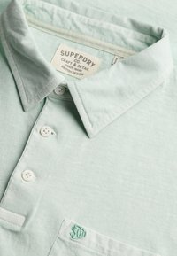 Polo Superdry vert clair avec trois boutons blancs, un col et un petit logo brodé sur la poche poitrine.