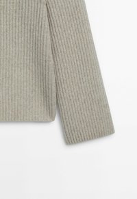 Manche de pull en maille côtelée beige avec un motif doux et texturé, présentée sur un fond blanc.