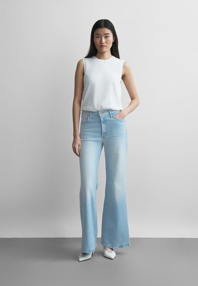 Νεαρή γυναίκα που στέκεται, φοράει λευκό αμάνικο τοπ, ανοιχτό μπλε τζιν με ψηλή μέση και φαρδιά παντελόνια, και λευκά παπούτσια με μυτερή μύτη, μπροστά σε απλό φόντο.