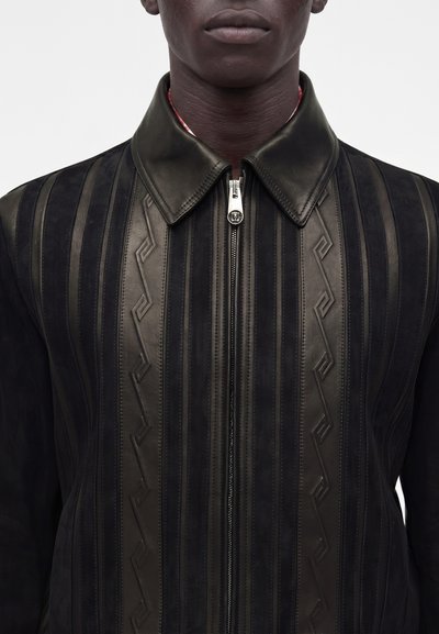Homme portant une veste en cuir noire avec des rayures verticales matelassées et des motifs géométriques, fermeture éclair fermée, fond uni.
