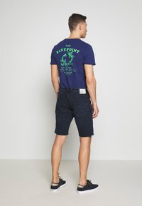 Marineblauwe t-shirt met groene graphic op de achterkant, gecombineerd met donkere shorts en zwarte schoenen. Eenvoudig ontwerp met een relaxed fit en geen zichtbare patronen.