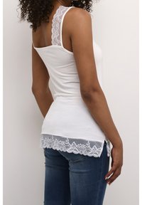 Camiseta sin mangas blanca con detalles de encaje en los hombros y el dobladillo. La tela es suave y ajustada, combinada con unos vaqueros de mezclilla azul.