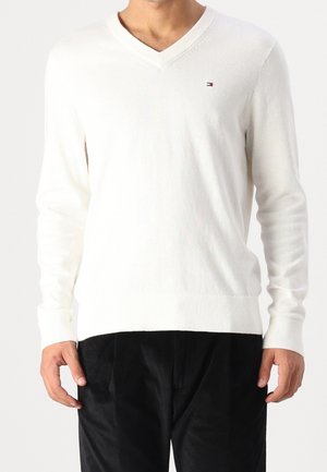Pull blanc à manches longues avec col en V et petit logo sur la poitrine, porté par une personne portant un pantalon noir, debout devant un fond uni.