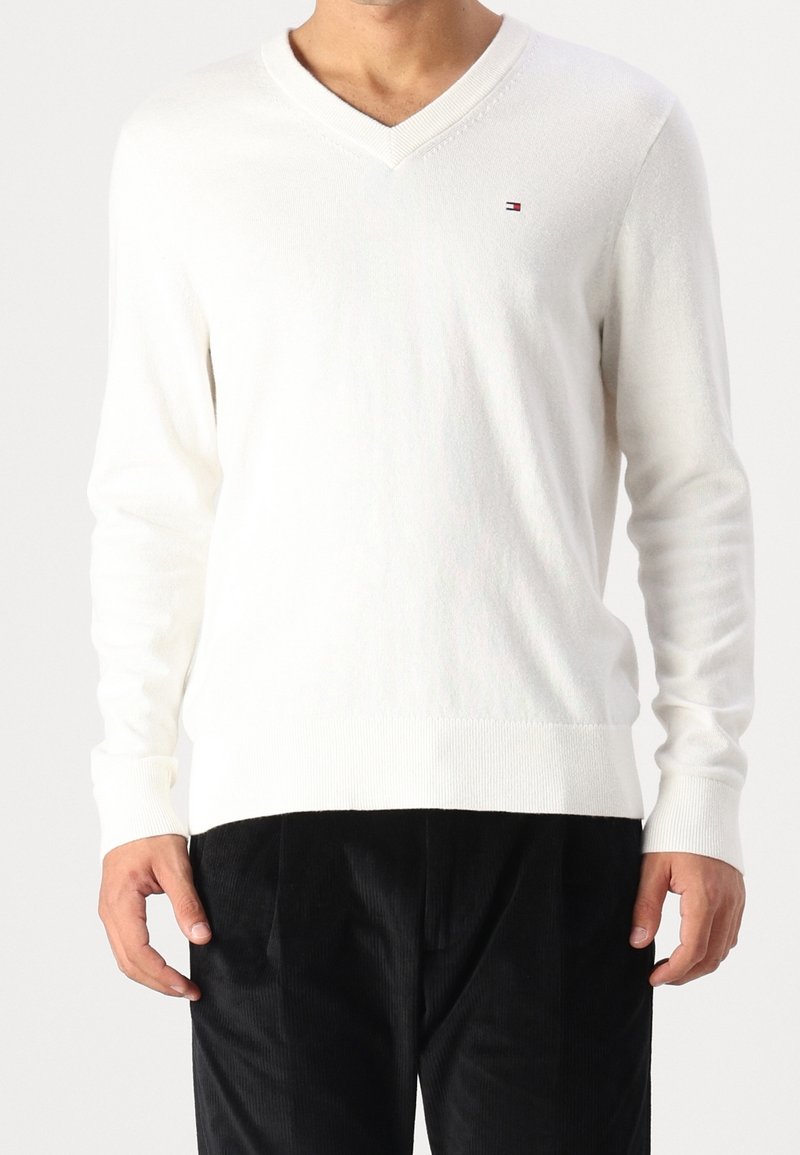 Pull blanc à manches longues avec col en V et petit logo sur la poitrine, porté par une personne portant un pantalon noir, debout devant un fond uni.
