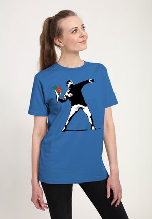 Frau trägt ein blaues Hemd mit einem Motiv einer Person, die einen Strauß roter Blumen wirft, posiert, als würde sie einen Molotow-Cocktail werfen.