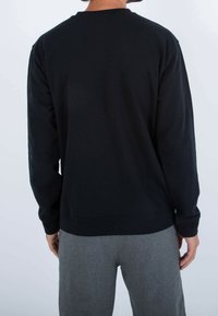 Osoba oblečená v jednoduchém černém tričku s dlouhým rukávem a šedých kalhotách, zobrazená ze zad na bílém pozadí.