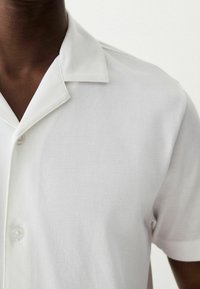 Weißes kurzärmeliges Hemd mit strukturierter Oberfläche, ausgestattet mit einem klassischen Kragen und einem einzelnen Knopfverschluss am Hals.